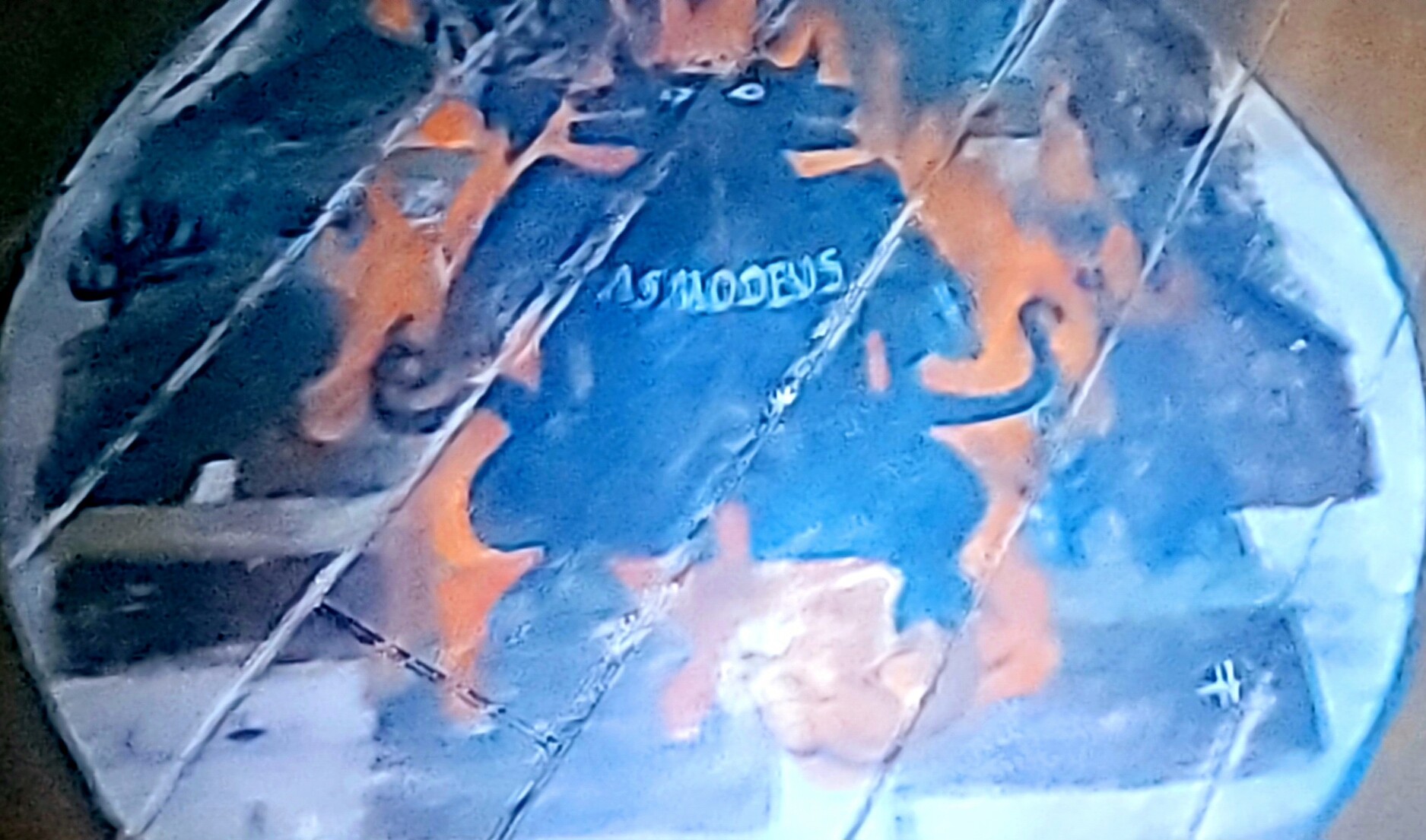 Screenshot from Paranormal Activity: Next of Kin showing a silhouette of a two headed goat-like monster surrounded by orange flames and black smudges in a white circle painted on the floor. The word ASMODEUS has been painted within the silhouette.